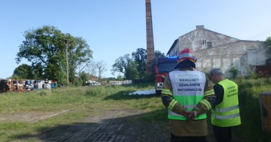 Inspektor Ochrony Środowiska wraz ze strażakiem w trakcie czynności służbowych. W tle wóz strażacki, budynki przemysłowe oraz odpady w beczkach i mauzerach.